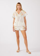 [Color: Cream/Coral] A front facing image of a blonde model wearing a cotton cream top with contrasting floral embroidered details. This relaxed cotton top features short sleeves and a split v-neckline with ties. Perfect for lounging or vacations. 