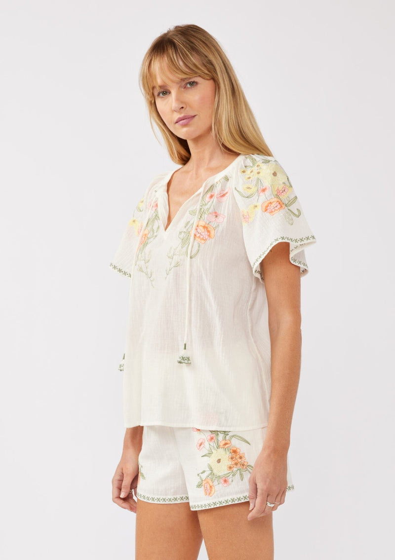 [Color: Cream/Coral] A side facing image of a blonde model wearing a cotton cream top with contrasting floral embroidered details. This relaxed cotton top features short sleeves and a split v-neckline with ties. Perfect for lounging or vacations. 