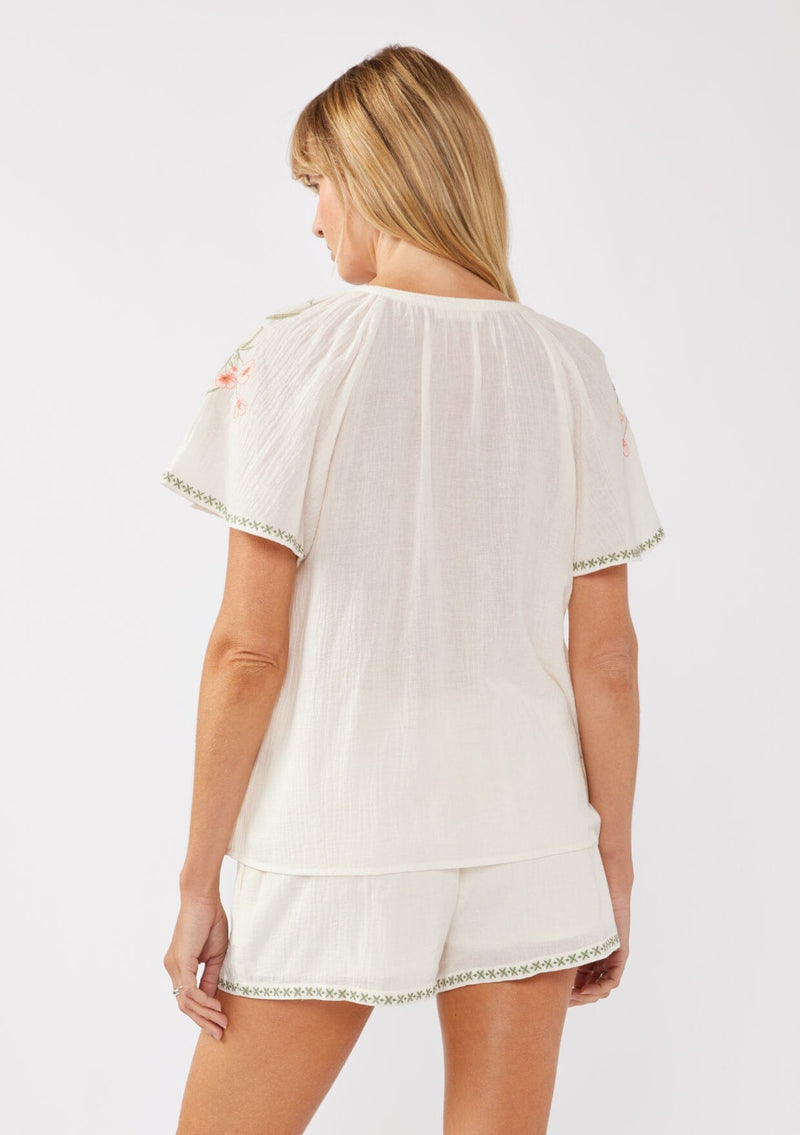 [Color: Cream/Coral] A back facing image of a blonde model wearing a cotton cream top with contrasting floral embroidered details. This relaxed cotton top features short sleeves and a split v-neckline with ties. Perfect for lounging or vacations. 