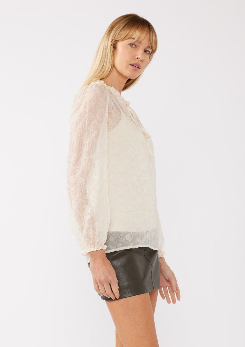 [Color: Ivory] A full body side facing image of a blonde model wearing a crinkled sheer chiffon blouse with a floral pattern in ivory off white . This relaxed fit blouse features a double tie neck detail, long sleeves with ruffled elastic cuffs, and a detachable cami. 