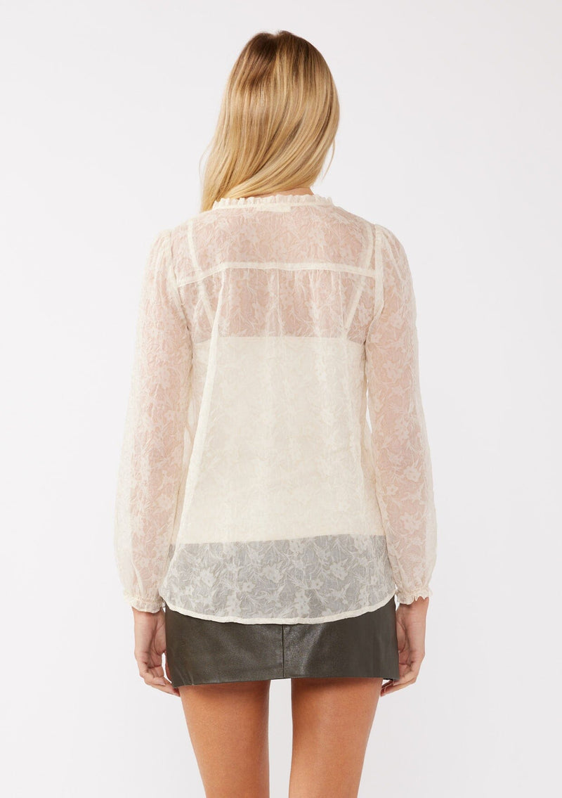 [Color: Ivory] A back facing image of a blonde model wearing a crinkled sheer chiffon blouse with a floral pattern in ivory off white . This relaxed fit blouse features a double tie neck detail, long sleeves with ruffled elastic cuffs, and a detachable cami. 