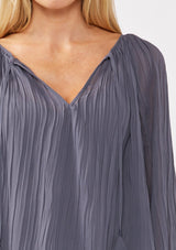 [Color: Slate Blue] A detail image of a blonde model wearing a blue pleated chiffon blouse. Featuring a split v-neckline with tie detail, long sleeves, and elastic cuffs. A flowy, relaxed top for the office or special occasions. 