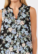 [Color: Black/Pale Blue] A detail image of a blonde model wearing a black and blue floral print blouse sleeveless top. Featuring a button front and a split v neckline with a ruffle trim detail. Perfect for work and special occasions. 