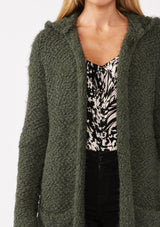 [Color: Olive] A detail image of a blonde model wearing a fuzzy army green fall cardigan with a popcorn texture. The cardigan features an open front, large cozy patch pockets, and an attached hoodie, draped for warmth and ideal for layering. 