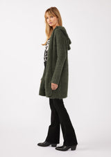 [Color: Olive] A side facing image of a blonde model wearing a fuzzy army green fall cardigan with a popcorn texture. The cardigan features an open front, large cozy patch pockets, and an attached hoodie, draped for warmth and ideal for layering. 