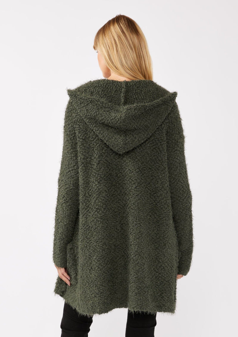 [Color: Olive] A back facing image of a blonde model wearing a fuzzy army green fall cardigan with a popcorn texture. The cardigan features an open front, large cozy patch pockets, and an attached hoodie, draped for warmth and ideal for layering. 