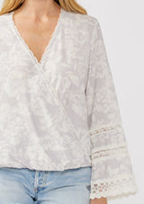 [Color: Dove/Lavender] A detail image of a blonde model wearing a purple floral print surplice top with lace trim details. This boho blouse features a flattering v-neckline, long bell sleeves, and a front elastic hem with a high low silhouette. Perfect for casual outings and resort styling. 