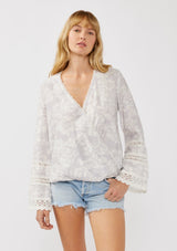 [Color: Dove/Lavender] A front facing image of a blonde model wearing a purple floral print surplice top with lace trim details. This boho blouse features a flattering v-neckline, long bell sleeves, and a front elastic hem with a high low silhouette. Perfect for casual outings and resort styling. 