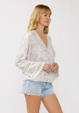 [Color: Dove/Lavender] A side facing image of a blonde model wearing a purple floral print surplice top with lace trim details. This boho blouse features a flattering v-neckline, long bell sleeves, and a front elastic hem with a high low silhouette. Perfect for casual outings and resort styling. 