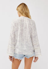 [Color: Dove/Lavender] A back facing image of a blonde model wearing a purple floral print surplice top with lace trim details. This boho blouse features a flattering v-neckline, long bell sleeves, and a front elastic hem with a high low silhouette. Perfect for casual outings and resort styling. 