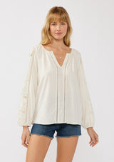 [Color: Natural] A front facing image of a blonde model wearing a rayon-linen blend blouse featuring a split V-neckline, long sleeves with embroidered cutout details, and elastic cuffs, styled tucked into denim shorts for a relaxed, resort-inspired look.