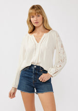 [Color: Natural] A front facing image of a blonde model wearing a rayon-linen blend blouse featuring a split V-neckline, long sleeves with embroidered cutout details, and elastic cuffs, styled tucked into denim shorts for a relaxed, resort-inspired look.