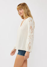 [Color: Natural] A side facing image of a blonde model wearing a rayon-linen blend blouse featuring a split V-neckline, long sleeves with embroidered cutout details, and elastic cuffs, styled tucked into denim shorts for a relaxed, resort-inspired look.