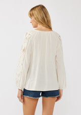 [Color: Natural] A back facing image of a blonde model wearing a rayon-linen blend blouse featuring a split V-neckline, long sleeves with embroidered cutout details, and elastic cuffs, styled tucked into denim shorts for a relaxed, resort-inspired look.