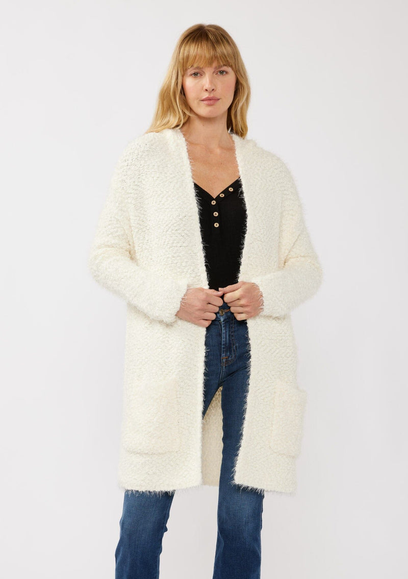 [Color: Antique White] A front facing image of a blonde model wearing a fuzzy white fall cardigan with a popcorn texture. The cardigan features an open front, large cozy patch pockets, and an attached hoodie, draped for warmth and ideal for layering. 