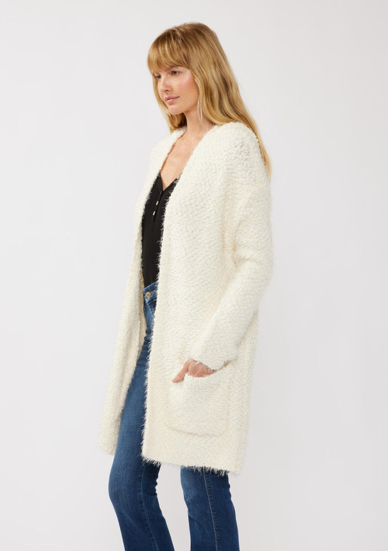 [Color: Antique White] A side facing image of a blonde model wearing a fuzzy white fall cardigan with a popcorn texture. The cardigan features an open front, large cozy patch pockets, and an attached hoodie, draped for warmth and ideal for layering. 