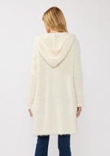 [Color: Antique White] A back facing image of a blonde model wearing a fuzzy white fall cardigan with a popcorn texture. The cardigan features an open front, large cozy patch pockets, and an attached hoodie, draped for warmth and ideal for layering. 