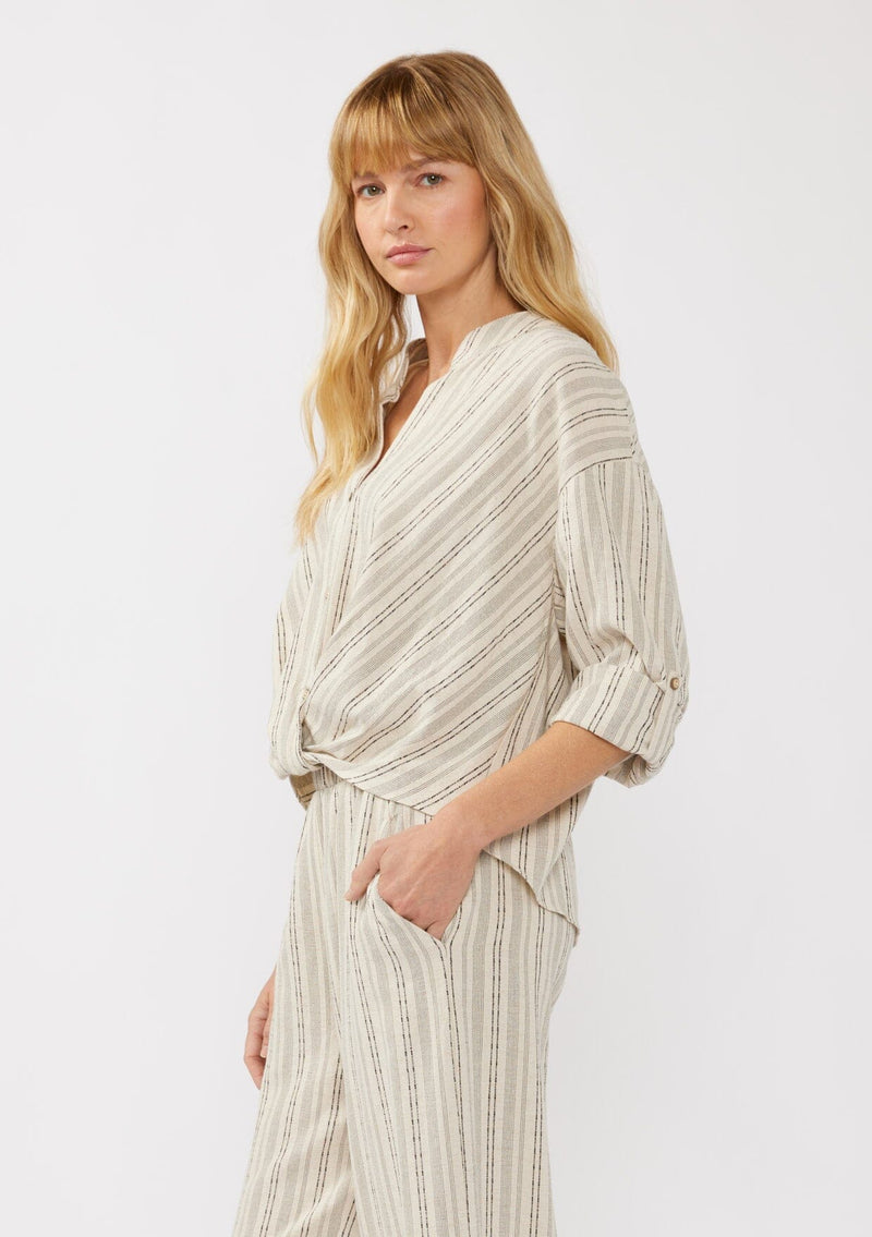 [Color: Natural/Black] A side facing image of a blonde model wearing a cotton blend blouse in beige with black yarn-dye stripes. The top features a split V-neckline, button front, front knot, and long sleeves with button tabs, ideal for lounging or vacation style.