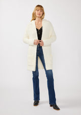 [Color: Antique White] A full body front facing image of a blonde model wearing a fuzzy white fall cardigan with a popcorn texture. The cardigan features an open front, large cozy patch pockets, and an attached hoodie, draped for warmth and ideal for layering. 