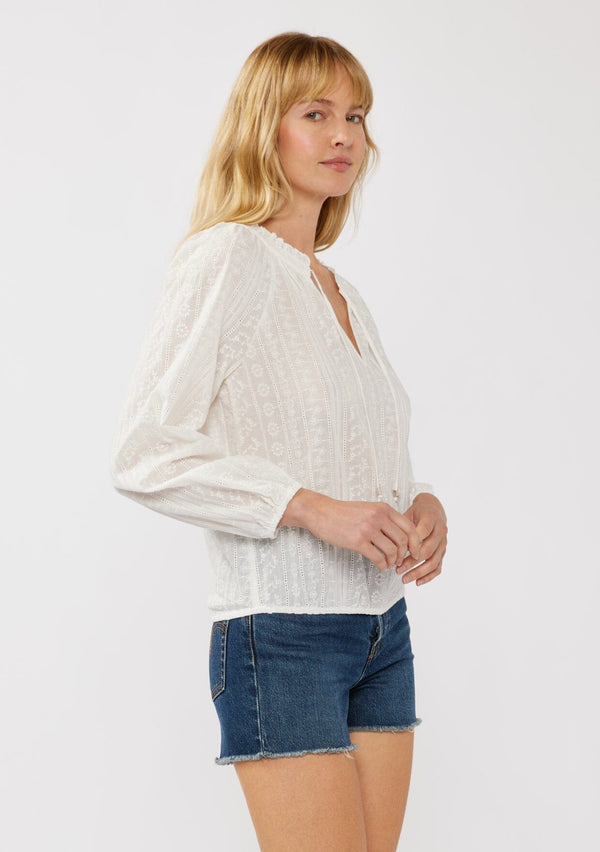 [Color: White] A side facing image of a blonde model wearing a white cotton top with floral embroidery. This sheer top features a split v-neckline with ties, long sleeves with elastic cuffs, and an elastic hemline.