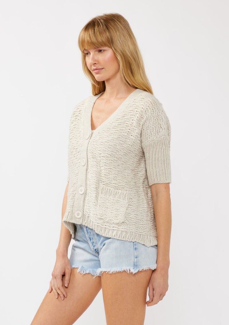 [Color: Bone] A side-facing image of a blonde model wearing a cozy, cropped knit cardigan with a V-neckline, button front, and ribbed half sleeves. Ideal for lounging or layering for a relaxed, casual style.