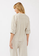 [Color: Natural/Black] A back facing image of a blonde model wearing a cotton blend blouse in beige with black yarn-dye stripes. The top features a split V-neckline, button front, front knot, and long sleeves with button tabs, ideal for lounging or vacation style.