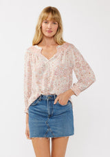 [Color: Natural/Dusty Pink] A front facing image of a blond model wearing a classic v-neck bohemian blouse with a pink abstract print, smocked neckline and cuffs, and button front. 