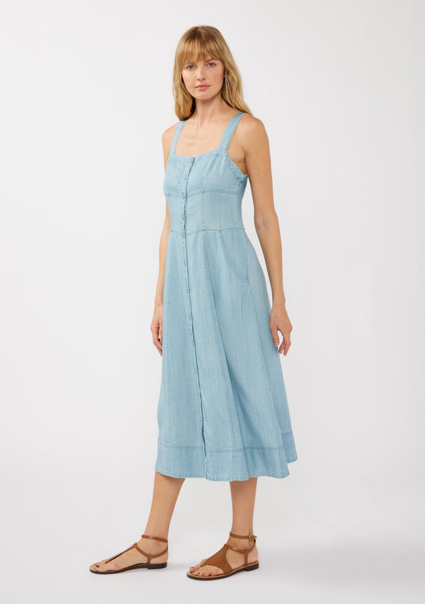 [Color: Light Wash] A side facing image of a blonde model in a blue denim-like Tencel midi dress, showcasing a square neckline, adjustable button-tab straps, button-front design, and handy side pockets—ideal for casual strolls, vacation days, or refined resort style.