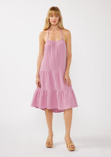 [Color: Orchid] A front facing image of a blonde model wearing a sleeveless halter mid length dress in a light purple cotton gauze. With an adjustable halter neckline, a tiered silhouette, a low back with adjustable tie closure, and a scooped neckline. 