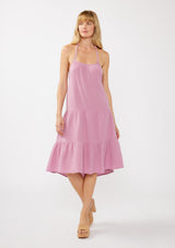 [Color: Orchid] A front facing image of a blonde model wearing a sleeveless halter mid length dress in a light purple cotton gauze. With an adjustable halter neckline, a tiered silhouette, a low back with adjustable tie closure, and a scooped neckline. 