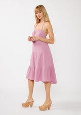 [Color: Orchid] A side facing image of a blonde model wearing a sleeveless halter mid length dress in a light purple cotton gauze. With an adjustable halter neckline, a tiered silhouette, a low back with adjustable tie closure, and a scooped neckline. 