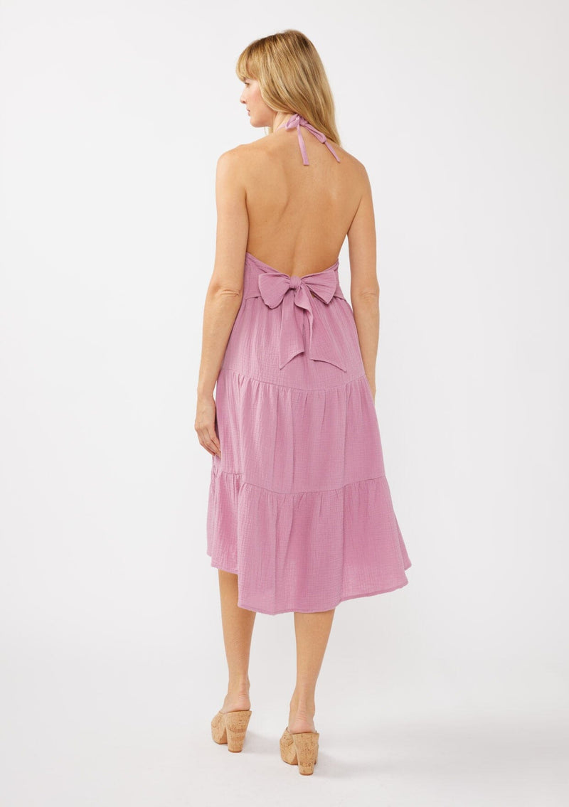 [Color: Orchid] A back facing image of a blonde model wearing a sleeveless halter mid length dress in a light purple cotton gauze. With an adjustable halter neckline, a tiered silhouette, a low back with adjustable tie closure, and a scooped neckline. 