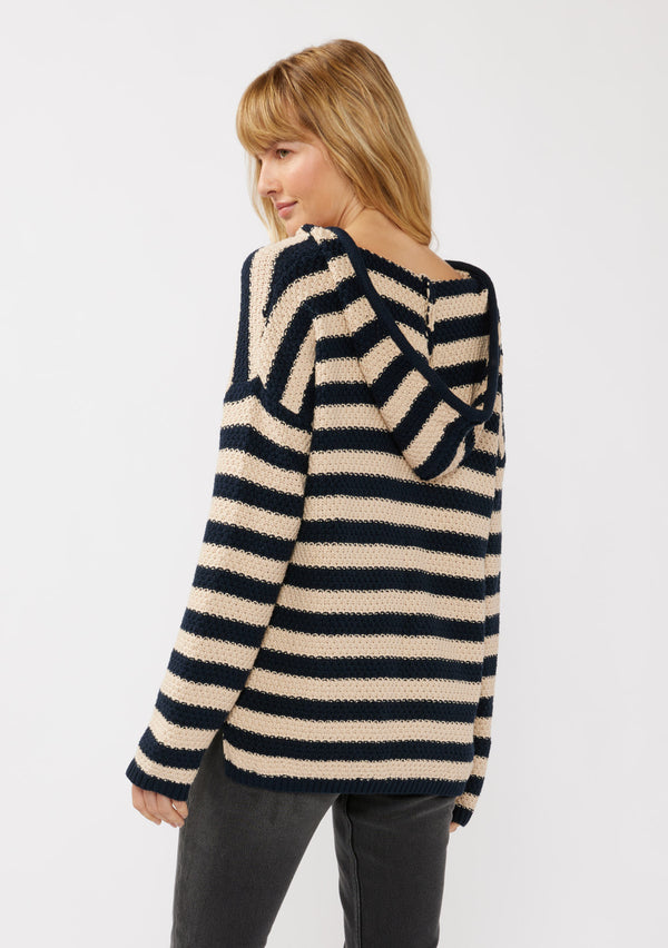 [Color: Navy/Natural] A side facing image of a blonde model wearing a navy and beige striped knit hoodie. With long sleeves, a dropped shoulder, a v neckline, and a front kangaroo pocket. 