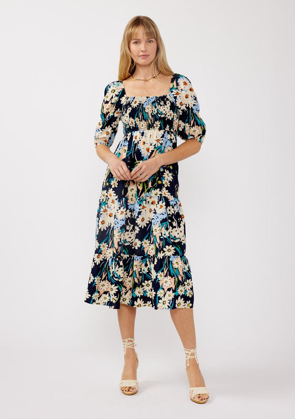 [Color: Navy/Natural] A front facing image of a blonde model wearing a navy square neck midi dress with a daisy floral print.  Featuring short puff sleeves with an elastic tiered detail, smocked bodice, and a tiered flowy skirt. Perfect for resort styling, casual outings, or brunch dates. 