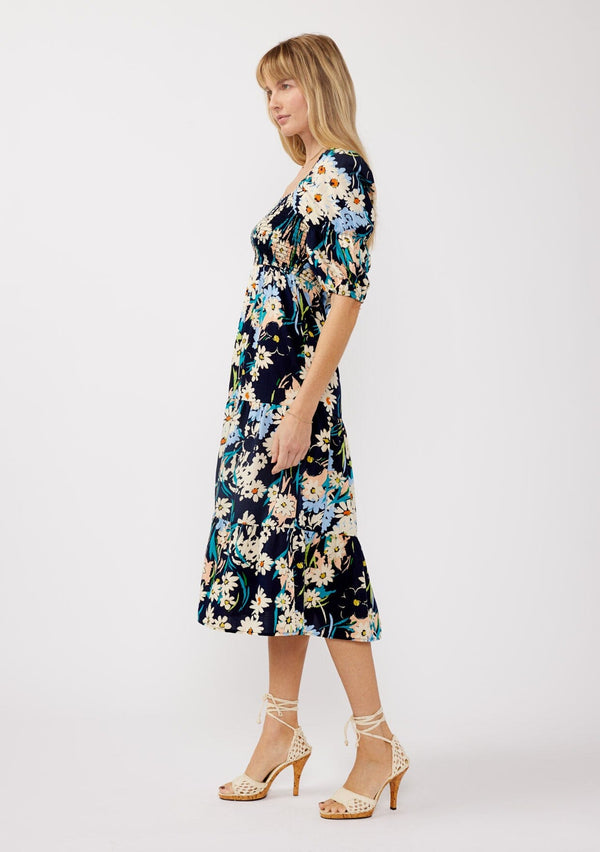 [Color: Navy/Natural] A side facing image of a blonde model wearing a navy square neck midi dress with a daisy floral print.  Featuring short puff sleeves with an elastic tiered detail, smocked bodice, and a tiered flowy skirt. Perfect for resort styling, casual outings, or brunch dates. 