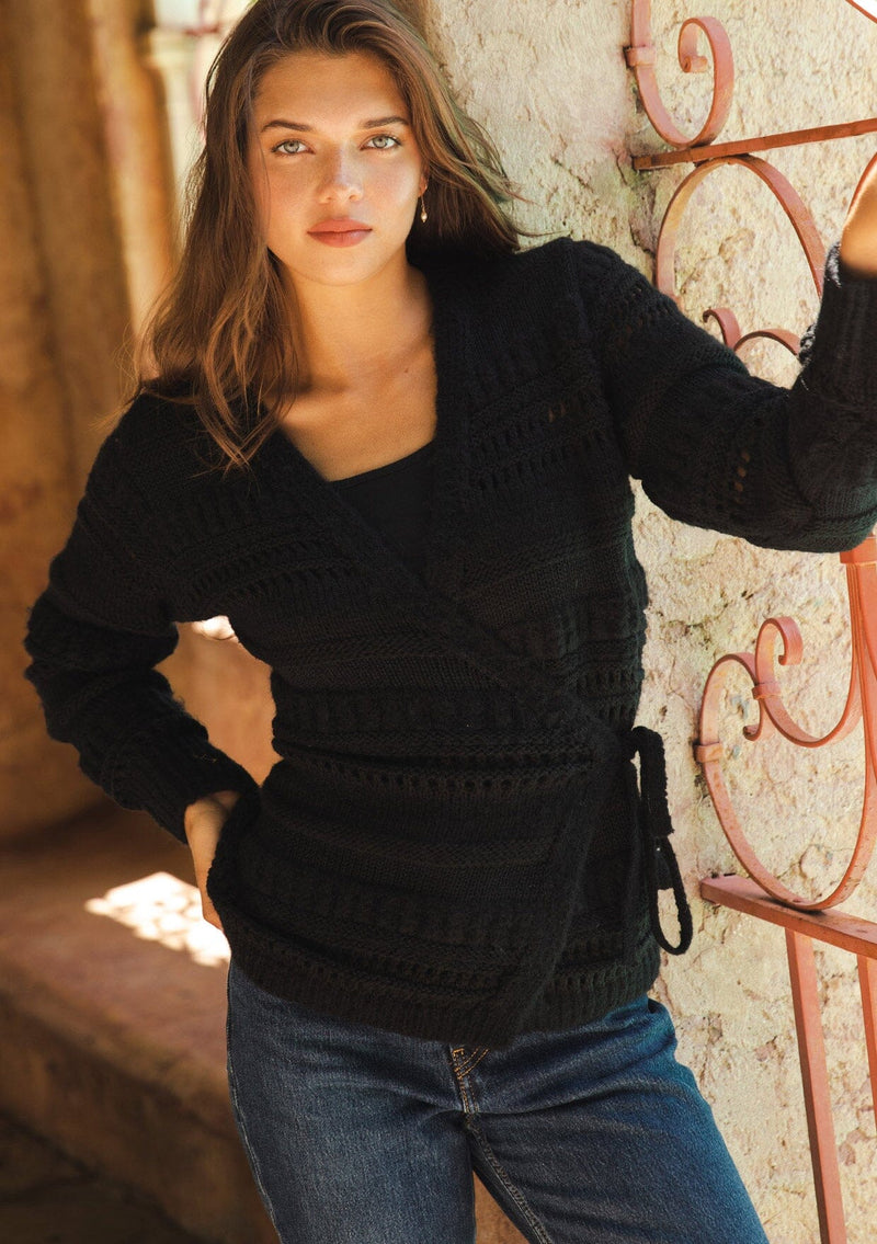 [Color: Black] A brunette model wearing a black sweater knit top with a wrap front and side tie closure. A chunky knit sweater with a mixed yarn weave and features an open knit design with a v neckline and long sleeves. 