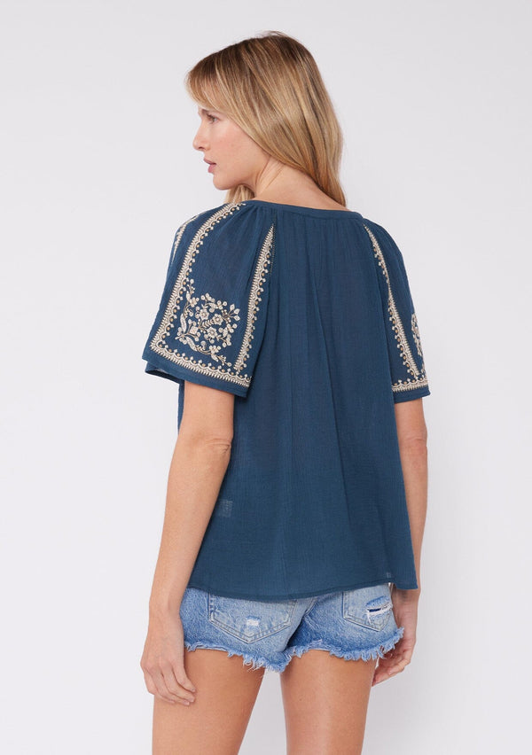 [Color: Teal/Natural] A back facing image of a blonde model wearing a teal blue cotton top with bohemian embroidered detail. With short raglan sleeves, a split v neckline with ties, and a relaxed fit. 