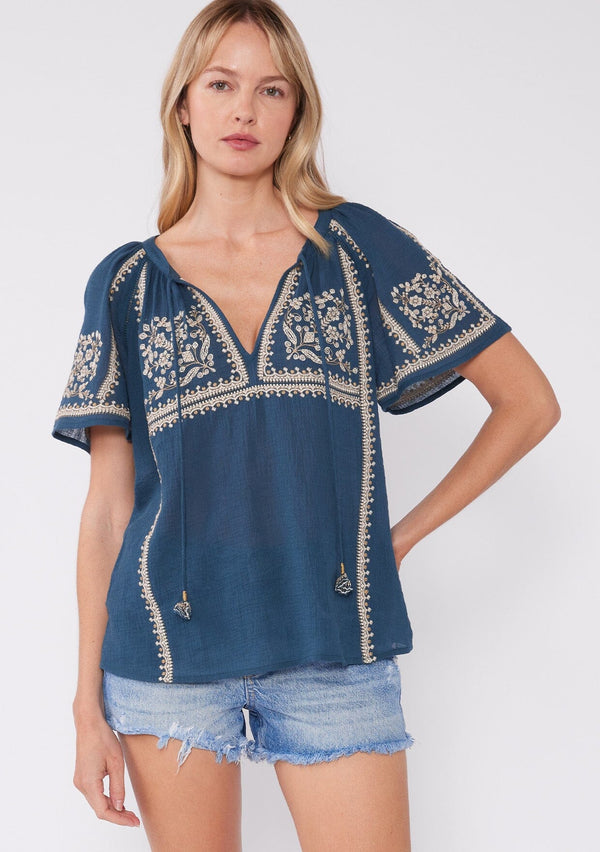 [Color: Teal/Natural] A front facing image of a blonde model wearing a teal blue cotton top with bohemian embroidered detail. With short raglan sleeves, a split v neckline with ties, and a relaxed fit. 