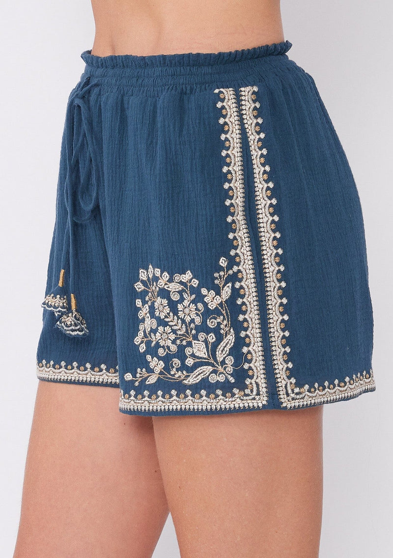 [Color: Teal/Natural] A close up side facing image of a blonde model wearing a teal blue bohemian cotton short with embroidered detail. With a smocked elastic waist, a drawstring tie waist, and side pockets. 