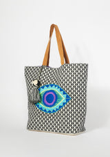 [Color: Black/Purple] An oversized bohemian tote bag with a black patterned exterior and contrast purple interior. With gold metallic lurex thread details, an embroidered evil eye motif, an oversized tassel accent, two suede top handles, and a magnetic snap closure. 
