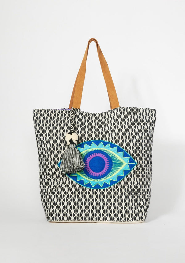 [Color: Black/Purple] An oversized bohemian tote bag with a black patterned exterior and contrast purple interior. With gold metallic lurex thread details, an embroidered evil eye motif, an oversized tassel accent, two suede top handles, and a magnetic snap closure. 