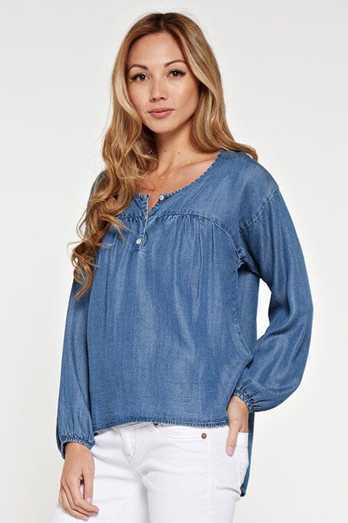 [Color: Vintage Wash] An asian model wearing a tencel blue denim blouse with a button front, yoke, long sleeves with dropped shoulders, and elastic wrist cuffs. 