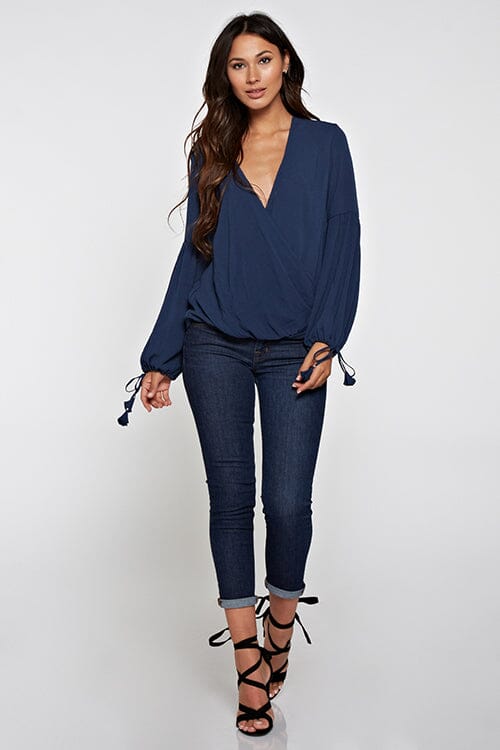 [Color: Deep Blue] A blonde model wearing a blue surplice v neckline blouse with long voluminous sleeves. Complete with a tassel tie detail on the cuff. Perfectly relaxed and ultra boho for any season. 