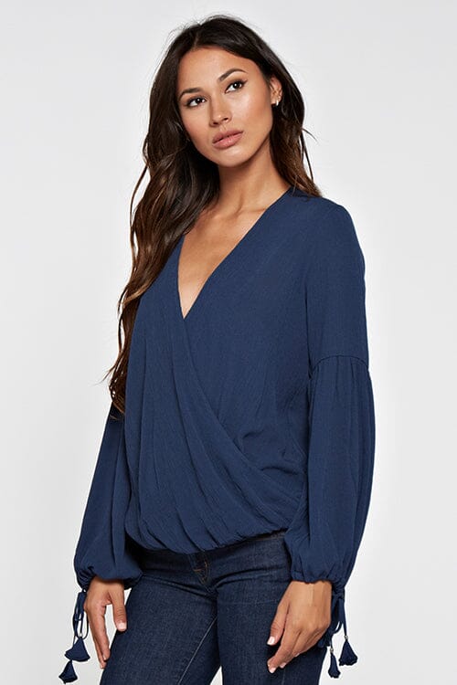 [Color: Deep Blue] A blonde model wearing a blue surplice v neckline blouse with long voluminous sleeves. Complete with a tassel tie detail on the cuff. Perfectly relaxed and ultra boho for any season. 