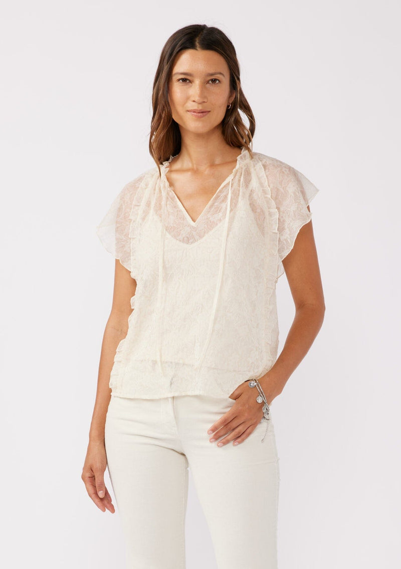 [Color: Ivory] A brunette model wearing a crinkled chiffon top with a floral pattern. This ivory off white top features a sheer flowy silhouette, flutter short sleeves, split v neckline with ties, and ruffled details with a merrow edge finish.  