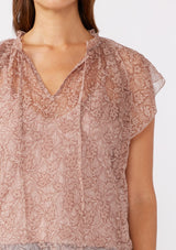 [Color: Mauve] A detailed image of a brunette model wearing a crinkled chiffon top with a floral pattern. This rose taupe top features a sheer flowy silhouette, flutter short sleeves, split v neckline with ties, and a ruffled details with a merrow edge finish.  