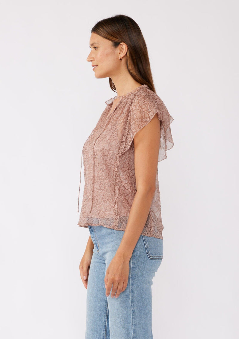 [Color: Mauve] A side image of a brunette model wearing a crinkled chiffon top with a floral pattern. This rose taupe top features a sheer flowy silhouette, flutter short sleeves, split v neckline with ties, and a ruffled details with a merrow edge finish.  