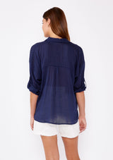 [Color: Navy] A brunette woman wearing a sheer blue shirt with a collared neckline, long roll tab sleeves, button front, and pintuck detaiils. A casual top paired with white shorts for the ultimate summer look.