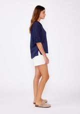 [Color: Navy] A brunette woman wearing a sheer blue shirt with a collared neckline, long roll tab sleeves, button front, and pintuck detaiils. A casual top paired with white shorts for the ultimate summer look.