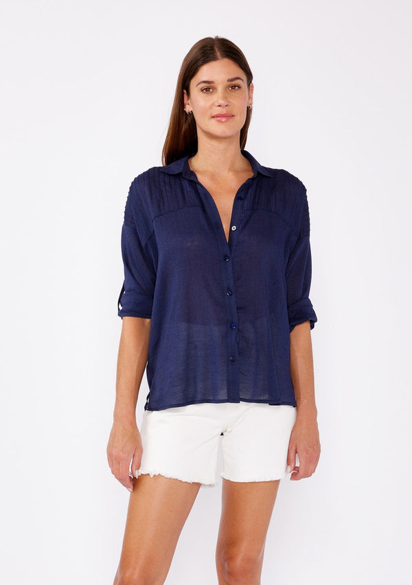 [Color: Navy] A brunette woman wearing a sheer blue shirt with a collared neckline, long roll tab sleeves, button front, and pintuck detaiils. A casual top paired with white shorts for the ultimate summer look. 
